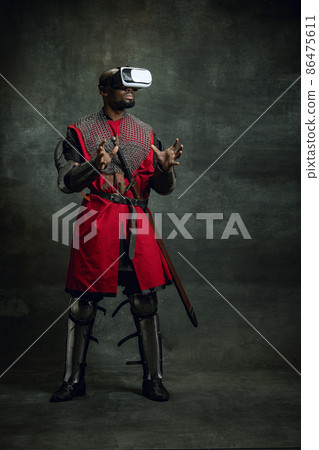Modern gadgets. One dark skinned man, medieval warrior or knight using VR headsets isolated over dark vintage background. Comparison of eras, history, technology 86475611