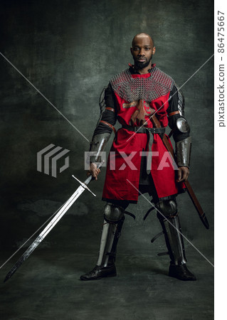 Vintage style portrait of brutal dark skinned man, medieval warrior or knight with wounded face wearing armour isolated over dark background. Comparison of eras, history Vintage style portrait of brutal dark skinned man, medieval warrior or knight with wounded face wearing armour isolated over dark background. Comparison of eras, history 86475667