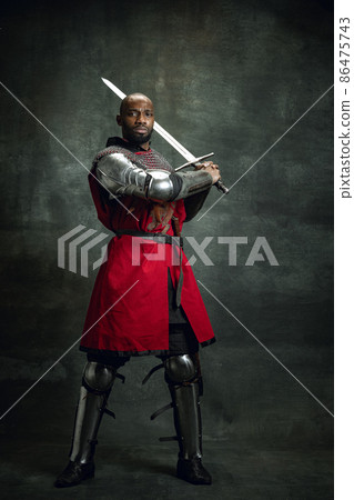 Vintage style portrait of brutal dark skinned man, medieval warrior or knight with wounded face wearing armour isolated over dark background. Comparison of eras, history 86475743