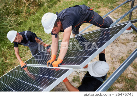 Male workers building photovoltaic solar panel system outdoors. Men engineers placing solar module on metal rails, wearing construction helmets and work gloves. Renewable and ecological energy. 86476745