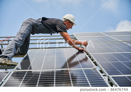 Worker mounting photovoltaic solar panel system outdoors. Man engineer placing solar module on metal rails, wearing construction helmets and work gloves. Renewable and ecological energy. Worker mounting photovoltaic solar panel system outdoors. Man engineer placing solar module on metal rails, wearing construction helmets and work gloves. Renewable and ecological energy. 86476749