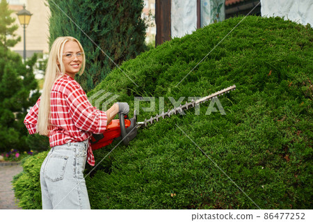 Young woman pruning bushes with electric hedge trimmer 86477252