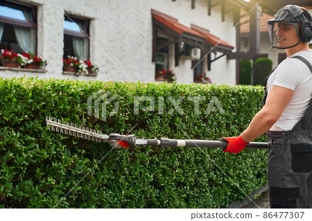 Man in uniform trimming bushes with hand electric cutter 86477307