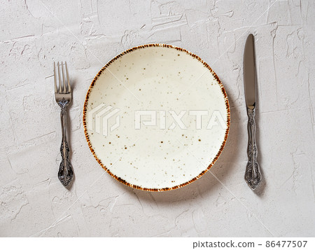 Set table. White speckled dinner plate near elegant table knife and fork over gray background. Set table. White speckled dinner plate near elegant table knife and fork over gray background. 86477507