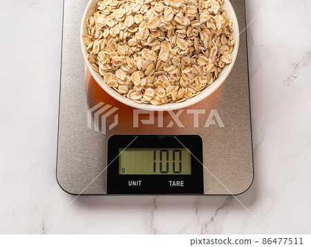 Raw dry oatmeal in a bowl on a digital kitchen scale. Raw dry oatmeal in a bowl on a digital kitchen scale. 86477511