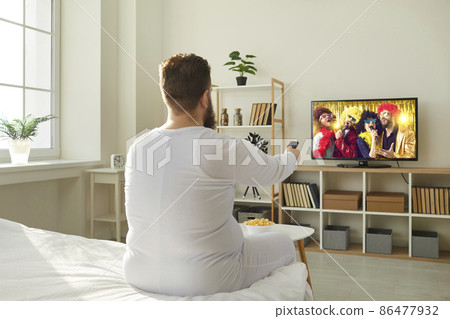 Plump ginger guy wakes up in morning and switches television with remote control. Funny fat man in PJs sitting in bedroom at home and watching entertainment channel on widescreen HD LED TV, back view 86477932