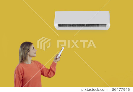 Happy woman switches on AC system. Young lady uses remote control to turn on AC on wall. Smiling girl standing isolated on yellow background setting comfortable temperature modern air conditioner Happy woman switches on AC system. Young lady uses remote control to turn on AC on wall. Smiling girl standing isolated on yellow background setting comfortable temperature modern air conditioner 86477946
