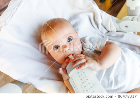 Cute little newborn girl drinking milk from bottle and looking at camera on white background. Infant baby sucking eating milk nutrition lying down on crib bed at home. Motherhood happy child concept Cute little newborn girl drinking milk from bottle and looking at camera on white background. Infant baby sucking eating milk nutrition lying down on crib bed at home. Motherhood happy child concept 86478317