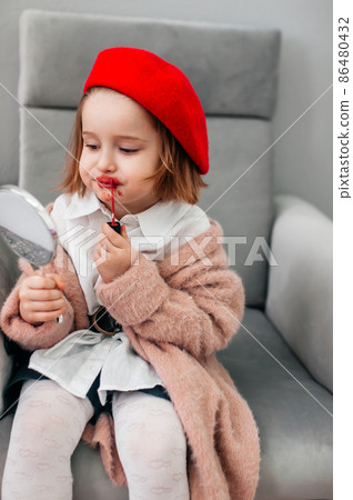 Red lipstick, bright baby lips. Fashionable child in a red beret and mother's shoes with heels. funny little girl making makeup with mother cosmetics Red lipstick, bright baby lips. Fashionable child in a red beret and mother's shoes with heels. funny little girl making makeup with mother cosmetics 86480432