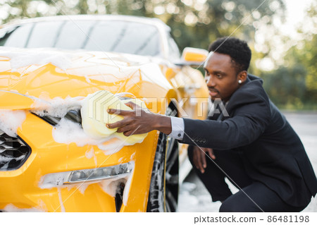 Young handsome bearded African businessman in formal wear cleaning his luxury sport car Young handsome bearded African businessman in formal wear cleaning his luxury sport car 86481198