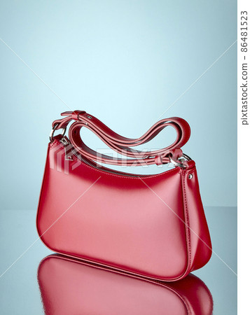 Fashionable handbag made of glossy red leather, standing on a mirror surface, on a pale blue background with a reflection. Copy space. 86481523