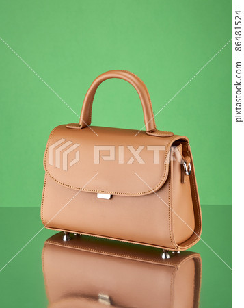 Fashionable handbag made of reddish-beige leather, standing on a mirror surface, on a rich green background with a reflection. Copy space. Fashionable handbag made of reddish-beige leather, standing on a mirror surface, on a rich green background with a reflection. Copy space. 86481524
