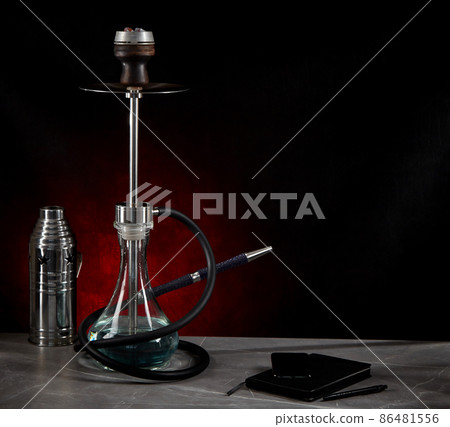 Beautiful modern hookah, stands on the table, surrounded by a metal protective cap and a diary, on a dark red background. Copy space. Beautiful modern hookah, stands on the table, surrounded by a metal protective cap and a diary, on a dark red background. Copy space. 86481556