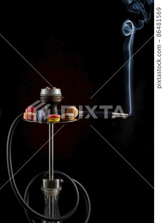Modern hookah with smoke rising from the mouthpiece, decorated with almond cookies of various colors on a dark background. Vertical image. The culture of serving hookah. Modern hookah with smoke rising from the mouthpiece, decorated with almond cookies of various colors on a dark background. Vertical image. The culture of serving hookah. 86481569