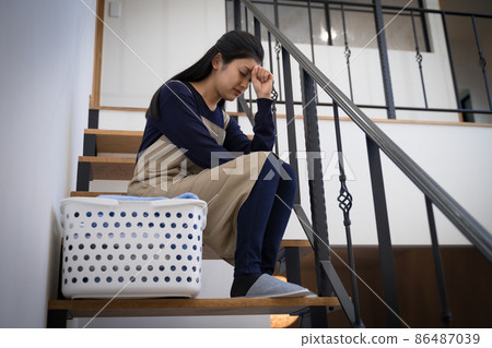 Image of a housewife tired of housework ・ A woman worried in front of a laundry basket on the stairs Image of a housewife tired of housework ・ A woman worried in front of a laundry basket on the stairs 86487039