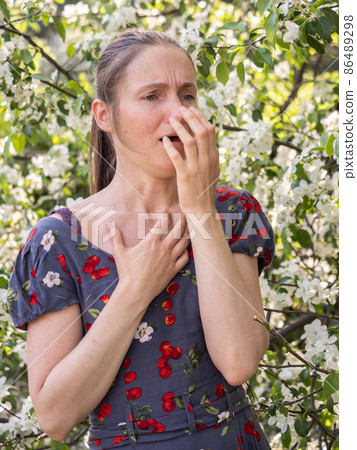Young woman sneezes in the park against the background of a flowering tree. Allergy to pollen concept. Young woman sneezes in the park against the background of a flowering tree. Allergy to pollen concept. 86489298