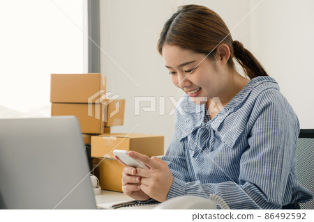 Young Asian woman business owner with many parcel boxes on the table happy online sales job, use the laptop, get an order from customers, take notes, and make arrangements for delivery by post. 86492592