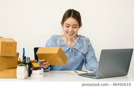Young Asian woman business owner with many parcel boxes on the table happy online sales job, use the laptop, get an order from customers, take notes, and make arrangements for delivery by post 86492619