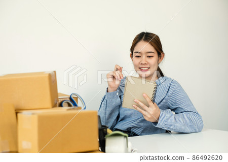 Young Asian woman business owner with many parcel boxes on the table happy online sales job, use the laptop, get an order from customers, take notes, and make arrangements for delivery by post 86492620