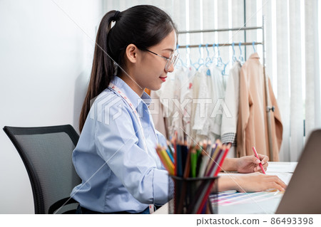 Beautiful young Asian fashion tailor with textile sewing accessories and entrepreneur designer sketches are full of bright colors on the desk with a laptop for creative ideas in the studio Beautiful young Asian fashion tailor with textile sewing accessories and entrepreneur designer sketches are full of bright colors on the desk with a laptop for creative ideas in the studio 86493398