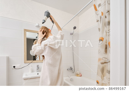 Back view of young woman wearing white bathrobe using hair dryer in bathroom Back view of young woman wearing white bathrobe using hair dryer in bathroom 86499167