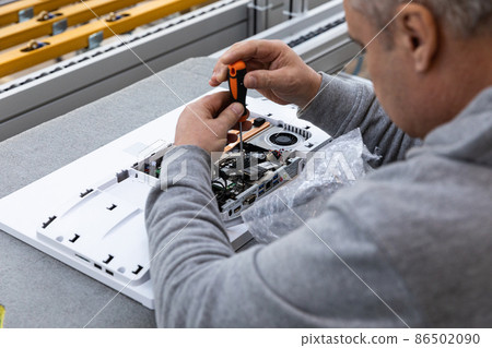 Photo of an adult man in a gray sweater who assembles a computer monitor system block on an conveyor 86502090