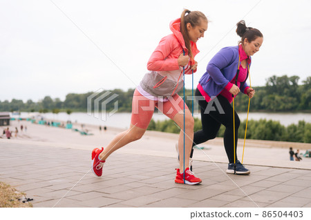 Side view of motivated fat young woman doing squats exercises using fitness tape for weight loss with personal trainer outdoor in summer day. Overweight female with coach stretching before running. Side view of motivated fat young woman doing squats exercises using fitness tape for weight loss with personal trainer outdoor in summer day. Overweight female with coach stretching before running. 86504403