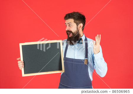 Surprised bearded man in barber apron hold school blackboard red background copy space, enrollment 86506965