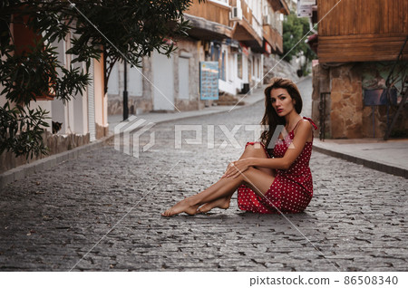 Caucasian girl in a red dress posing sitting on paving stone on a beautiful European street 86508340