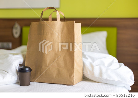 Delivery food craft bag with cup on the bed on sunny day. Delivery in any weather around the clock to the client. Fast food eco packaging with big breakfast set Delivery food craft bag with cup on the bed on sunny day. Delivery in any weather around the clock to the client. Fast food eco packaging with big breakfast set 86512210