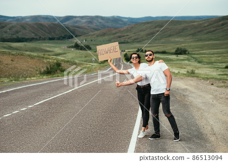 A beautiful couple is hitchhiking. A man and a woman are voting on the road. The couple tries to stop passing transport. People travel on their own. Travel around the country. Domestic tourism. A beautiful couple is hitchhiking. A man and a woman are voting on the road. The couple tries to stop passing transport. People travel on their own. Travel around the country. Domestic tourism. 86513094