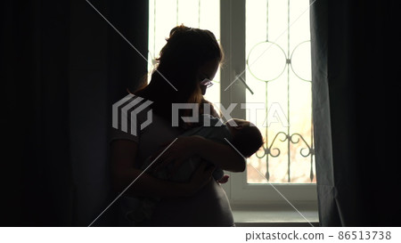 A young mother holds a child in her arms against the background of a window. The light from the window illuminates the image of a woman with a newborn baby. 86513738