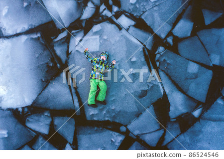 Little traveler kid boy in bright winter clothes lying on breaking ice in frozen water at river. Child playing on danger thin cracked ice. Top view of alone child boy in could water 86524546