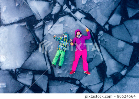 Woman and kid lie on the cracked ice of frozen river. Top view of travelers pose for a drone on an icy lake. Child and mother on block of ice in water. People in bright winter clothes 86524564