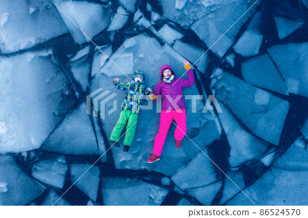 Couple lie on the transparent ice of the lake, view from above, aerial top view. Happy woman and kid traveller laying on the cracked ice on winter river. blocks of ice. People ib bright clothes Couple lie on the transparent ice of the lake, view from above, aerial top view. Happy woman and kid traveller laying on the cracked ice on winter river. blocks of ice. People ib bright clothes 86524570