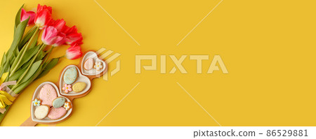 Sweets, pastry, gingerbread cookies for Easter table. Easter eggs heart shaped decor plate, pink tulips on yellow background top view copy space, spring seasonal holiday banner for site, flyer, coupon 86529881