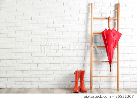 Beautiful red umbrella on ladder and rubber boots near white brick wall. Space for text Beautiful red umbrella on ladder and rubber boots near white brick wall. Space for text 86531307