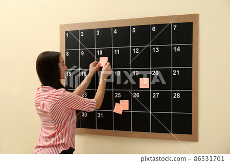 Young woman putting sticker on board calendar indoors Young woman putting sticker on board calendar indoors 86531701