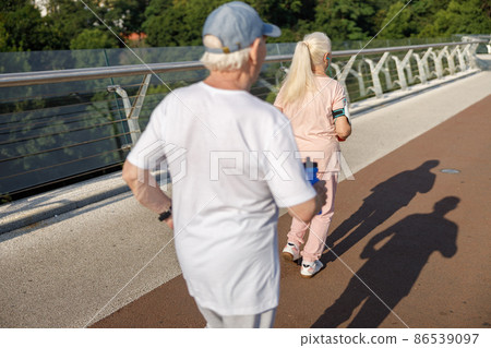Pair of active pensioners runs together along footbridge on summer day backside view Pair of active pensioners runs together along footbridge on summer day backside view 86539097