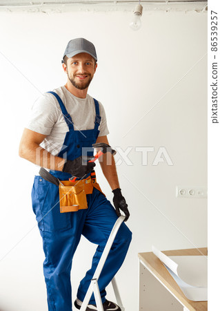 Cheerful electrician in uniform smiling at camera, holding hammer while installing light fitting in new apartment 86539257