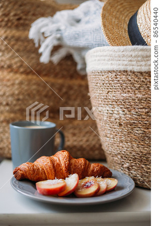 Freshly baked croissant and coffee mug. Coffee in cup on wooden table in cafe. Basket with a gray plaid and a hat 86540440