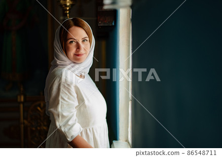 a woman in headscarf and light-colored robes by window in church.  86548711