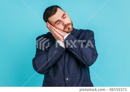 Bedtime. Portrait of handsome bearded man in suit sleeping laying down on her palms and smiling pleased, having comfortable nap and resting, dozing off. Indoor studio shot isolated on blue background. 86550371