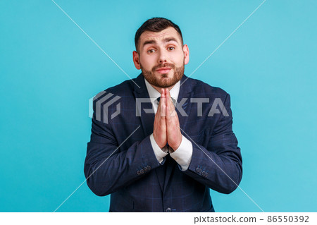 Please, I'm begging. Unhappy bearded man in official style suit keeping hands in prayer looking with imploring expression, sincere asking permission. Indoor studio shot isolated on blue background. 86550392