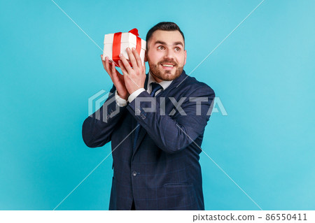 Curious dreamy bearded man in elegant suit shaking gift box trying to guess what is inside, wondered with present, surprises and bonuses. Indoor studio shot isolated on blue background. 86550411
