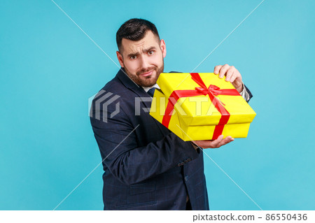 Portrait of upset curious man with beard holding open gift box, unpacking present to know what he received, frustrated and disappointed with content. Indoor studio shot isolated on blue background. 86550436
