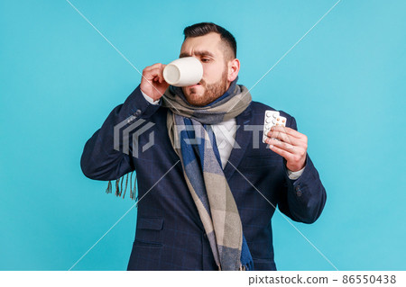 Sick businessman wearing official style suit and wrapped in scarf, holding pills in hands and drinking warm tea, suffering from fever or sore throat. Indoor studio shot isolated on blue background. Sick businessman wearing official style suit and wrapped in scarf, holding pills in hands and drinking warm tea, suffering from fever or sore throat. Indoor studio shot isolated on blue background. 86550438