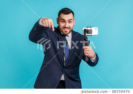 Businessman wearing official style suit, pointing down while communicating by video call or streaming, using steadicam, gesturing to subscribe. Indoor studio shot isolated on blue background. Businessman wearing official style suit, pointing down while communicating by video call or streaming, using steadicam, gesturing to subscribe. Indoor studio shot isolated on blue background. 86550470