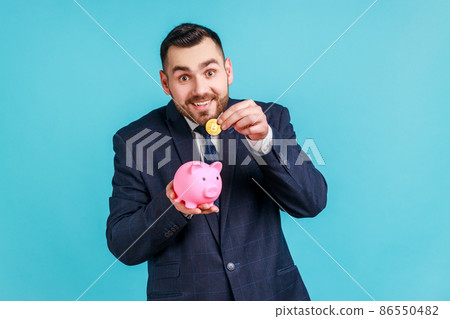 Attractive smiling young adult man with pleasant appearance, wearing suit holding piggy bank and putting bitcoin inside, happy with money savings. Indoor studio shot isolated on blue background. Attractive smiling young adult man with pleasant appearance, wearing suit holding piggy bank and putting bitcoin inside, happy with money savings. Indoor studio shot isolated on blue background. 86550482
