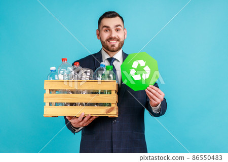 Satisfied man wearing official style suit holding box with plastic bottles and recycling green symbol, sorting his rubbish, saving ecology. Indoor studio shot isolated on blue background. Satisfied man wearing official style suit holding box with plastic bottles and recycling green symbol, sorting his rubbish, saving ecology. Indoor studio shot isolated on blue background. 86550483